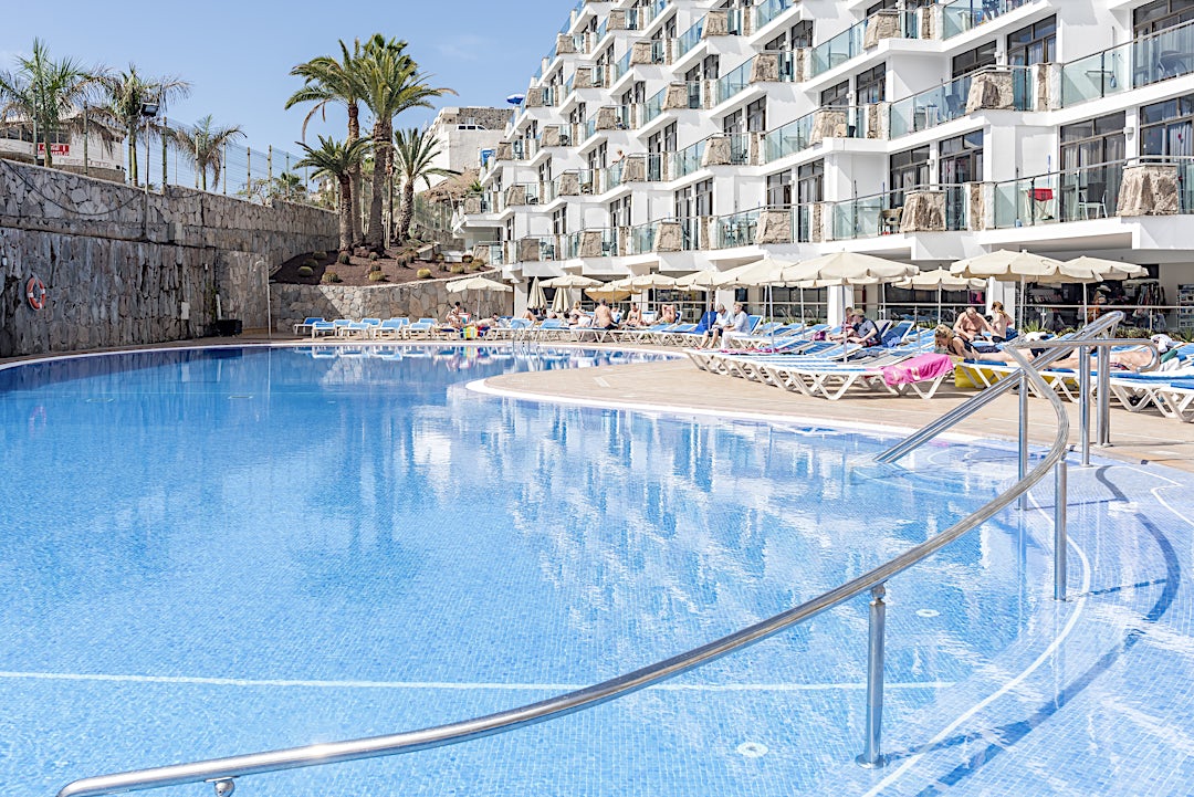 Poolområdet på Revoli Playa med liggestole og den hvide hotelbygning i baggrunden.