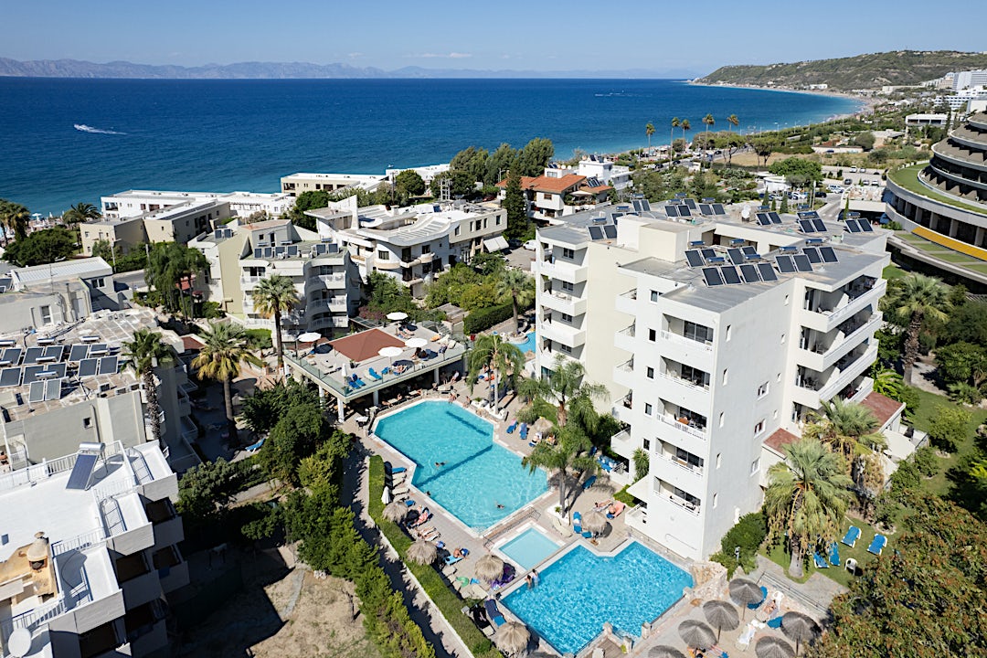 Hvid fleretagers hotelbygning med turkise swimmingpools på ejendommen med udsigt over kystby og blåt hav.