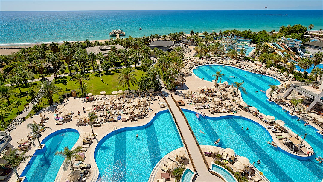 Flere turkisfarvede swimmingpools med snoede former, en lazy river og anlagte områder ved siden af en strand og hav.
