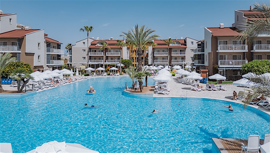 Stor resort swimmingpool med turkisblåt vand omgivet af hvide bygninger med terrakottatage og palmer i middelhavsstil.