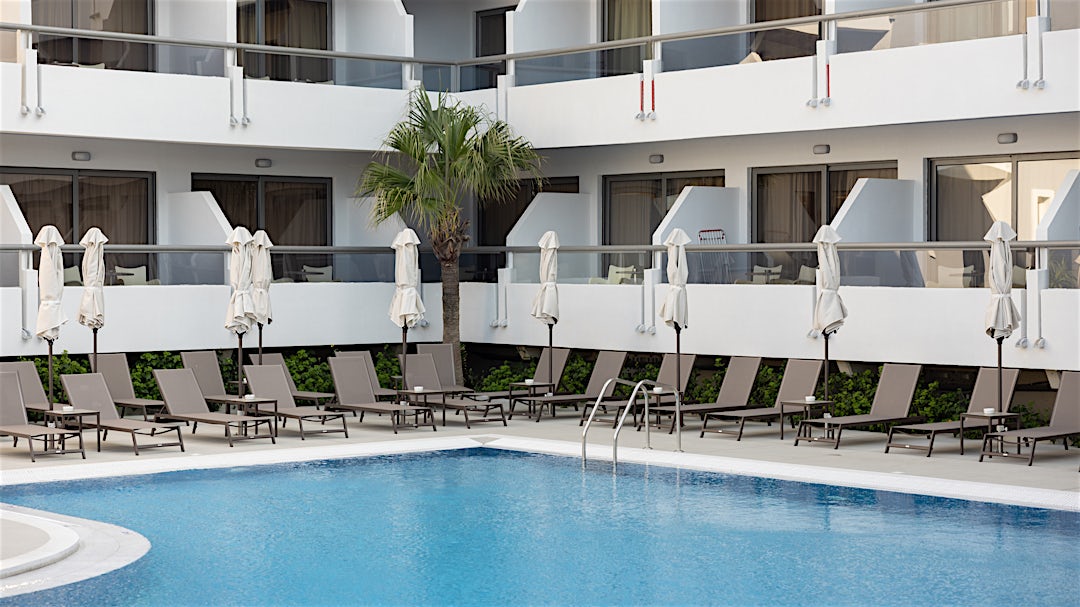 Poolområde med liggestole og hvid bygning i baggrunden.
