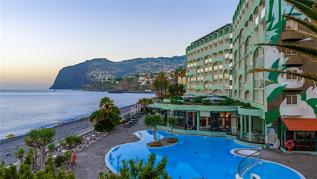 Hotelbygning med pool foran og udsigt over hav og bjerge i baggrunden.